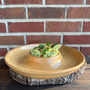 May include: A light brown ceramic serving dish with a central dip bowl, filled with guacamole and garnished with cilantro and lime wedges. The dish is resting on a wooden slice.