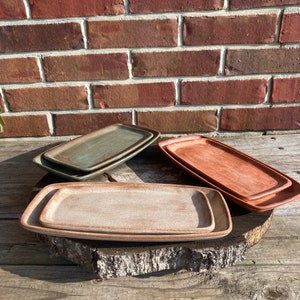 May include: Four rectangular ceramic serving trays in different shades of brown, green, and beige. The trays are stacked on top of each other on a wooden surface.