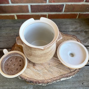 May include: A brown and white ceramic jar with a lid and a small strainer. The jar has a textured surface and is sitting on a wooden surface.