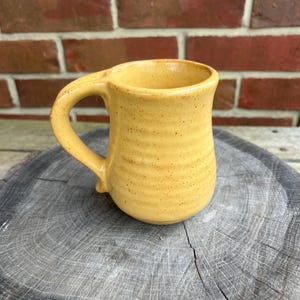 May include: A handmade yellow ceramic mug with a curved handle. The mug has a speckled texture and a slightly flared rim. The mug is sitting on a wooden surface, with a brick wall in the background.