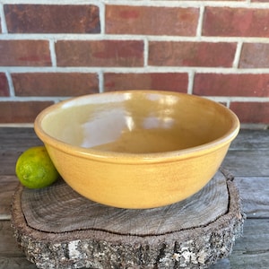 May include: A large, yellow ceramic bowl sits on a wooden slice. The bowl has a smooth, glossy finish and a slightly rounded shape.