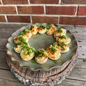 May include: A green ceramic plate with a scalloped edge holds a dozen deviled eggs topped with chopped green onions and a red sauce. The plate is on a wooden stump.