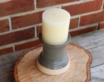 May include: A gray ceramic candle holder with a white base sits on a wooden slice. A white candle is in the holder.