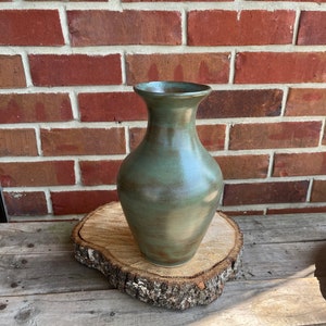 May include: A green ceramic vase with a speckled glaze. The vase has a rounded body and a narrow neck. It is sitting on a wooden slice.