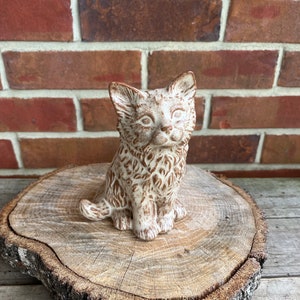 May include: A ceramic figurine of a brown and white cat sitting on a wooden slice. The cat has a detailed face and fur pattern.