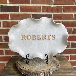 May include: A white ceramic tray with a scalloped edge and a speckled finish. The tray has the name "ROBERTS" engraved in brown lettering. The tray is sitting on a wooden stand.