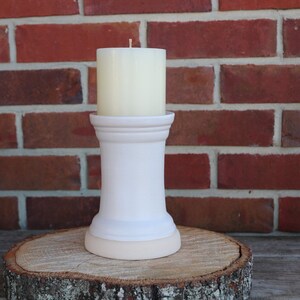 May include: White ceramic candle holder with a white pillar candle on top. The candle holder has a round base and a cylindrical body. The candle holder is sitting on a wooden surface.