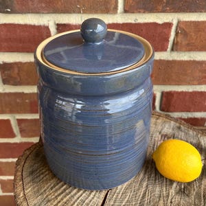 May include: A blue ceramic jar with a lid. The jar has a light brown rim and a subtle striped pattern. The jar is sitting on a wooden surface.