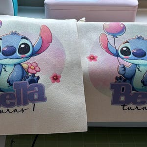 May include: Two white fabric bags with a blue Stitch character holding a pink balloon. The bags have the text "Bella turns 1" printed on them.