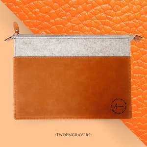 May include: A rectangular laptop sleeve with a color block design. The top section is light gray felt, the middle is a tan leather, and the bottom is a darker tan leather. The sleeve has a zipper closure and the word "Aurora" is printed on the bottom right.