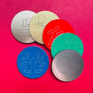 Pode incluir: Uma coleção de fichas metálicas redondas em várias cores, cada uma com um gráfico branco único. Os designs incluem uma fita, uma pessoa, uma figura correndo, uma pessoa reclinada, uma flecha atingindo um alvo e uma seta para cima com folhas.