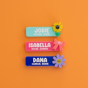 May include: Three rectangular name badges with white text. The top badge is light blue with the name JODIE and PATIENT FRIEND, featuring a yellow sunflower. The middle is pink with ISABELLA and SOCIAL WORKER, featuring a pink bow. The bottom is blue with DANA and CLINICAL NURSE, featuring a purple flower.