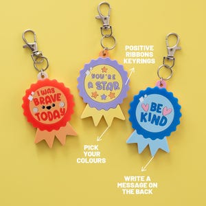 May include: Three circular award ribbon keychains with silver metal clasps. The red one says I WAS BRAVE TODAY, the purple one says YOU'RE A STAR, and the blue one says BE KIND. Text overlays include POSITIVE RIBBONS KEYRINGS, PICK YOUR COLOURS, and WRITE A MESSAGE ON THE BACK.