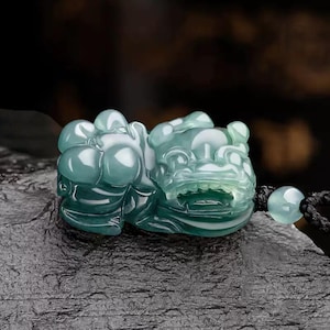 May include: A jade pendant carved in the shape of a mythical creature. The pendant is a translucent teal color with white veining. It is attached to a black cord with a matching teal bead.