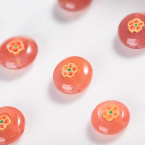 以下が含まれることがあります： 楕円形で半透明なピーチ色のビーズのクローズアップ。各ビーズには、緑色の中心を持つ金色の花が特徴です。ビーズは白い表面に散らばっており、視覚的に魅力的な配置になっています。