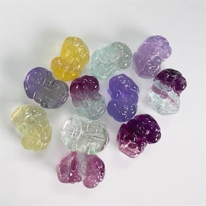 Puede incluir: Una colección de figuras talladas en piedras preciosas en varios colores, incluyendo amarillo, verde, morado y rosa. Las piedras translúcidas tienen forma de pequeñas figuras abstractas, dispuestas sobre una superficie blanca.