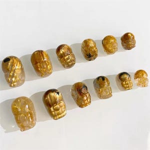 May include: A selection of golden crystal figurines, possibly owl-shaped, displayed on a white surface. The carvings have a translucent quality with internal golden inclusions, creating a shimmering effect. The figurines vary in size, with a range of detail.