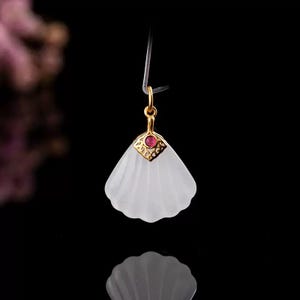 May include: A white, fan-shaped pendant with a gold-colored top and a small, red gemstone. The pendant is suspended by a thin, clear string against a black background. The design is elegant and minimalist.