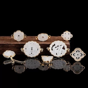 May include: A collection of white jade pendants and charms with gold-colored metal accents. The pendants feature various carved designs, including floral and geometric shapes. The items are displayed on a dark wooden surface against a black background.