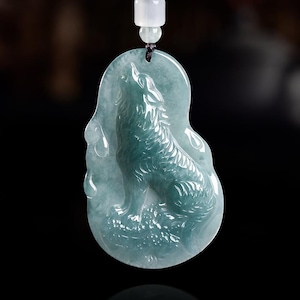 May include: A translucent, pale green jade pendant carved into an irregular oval shape featuring a detailed relief of a wolf howling. The pendant is suspended from a black cord with a small, cylindrical, white stone bead above the main piece.