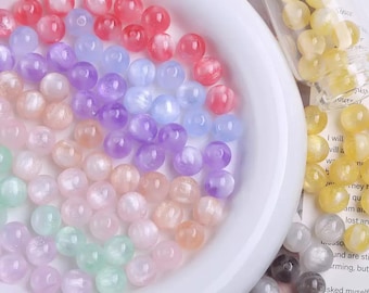 May include: A collection of colorful, translucent beads in a white bowl. The beads are in shades of red, purple, blue, green, and peach. Some beads are in a clear glass container. The beads are likely for jewelry making or crafts.