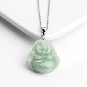 Puede incluir: Colgante de jade verde claro con forma de Buda sonriente, suspendido de una fina cadena de plata. La figura de Buda tiene una expresión serena y está detallada con curvas suaves. El colgante es un símbolo de buena suerte.