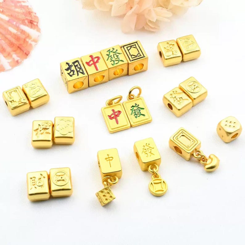 May include: Gold-colored charms and beads with Chinese characters and symbols. The collection includes cube-shaped beads, rectangular charms, and a single die. The characters are in red and green. The charms are likely for jewelry making.