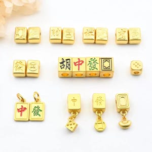 May include: Gold-colored Mahjong tile charms and beads. The charms feature Chinese characters in red and green. The beads have various designs and characters. Some charms have dangling dice or coin-shaped pendants. The items are arranged on a white surface.