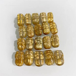 May include: Twenty golden-coloured, carved gemstone pendants are displayed. Each pendant has a barrel shape with detailed carvings. The gemstones are translucent, with visible internal structures. The pendants are arranged on a white surface.