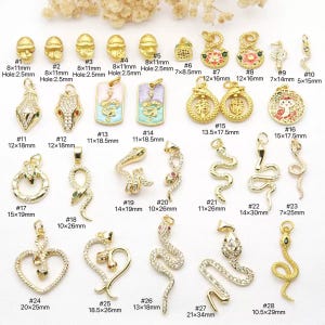 Op de afbeelding: Een verzameling goudkleurige sieradenbedels in diverse ontwerpen, waaronder slangen, harten en hangers. De bedels hebben afmetingen van 7x8,5 mm tot 21x34 mm. Sommige bedels zijn versierd met kristallen en gekleurd email.