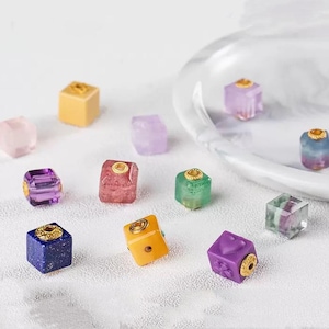 May include: A collection of colorful, cube-shaped beads in various colors, including pink, purple, yellow, green, and blue. Each bead has a gold-colored accent. The beads are scattered on a white surface, with some near a clear, round dish.