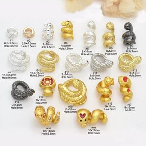Pode incluir: Uma coleção de contas metálicas em forma de cobra em ouro, prata e preto. As contas têm tamanhos diferentes, de 6,5 mm x 4,5 mm a 15 mm x 18 mm, com diferentes diâmetros de furo.
