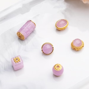 May include: An assortment of lavender-colored gemstone beads with gold-colored accents. The beads include a cylindrical bead, a cube-shaped bead, and several round and oval beads. The gold accents feature intricate designs, and the beads are arranged on a white surface.