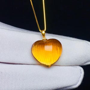 May include: A heart-shaped pendant with a golden hue, suspended from a delicate gold chain. The pendant appears to be made of a translucent material, catching the light and creating a warm glow. The necklace is set against a dark background.