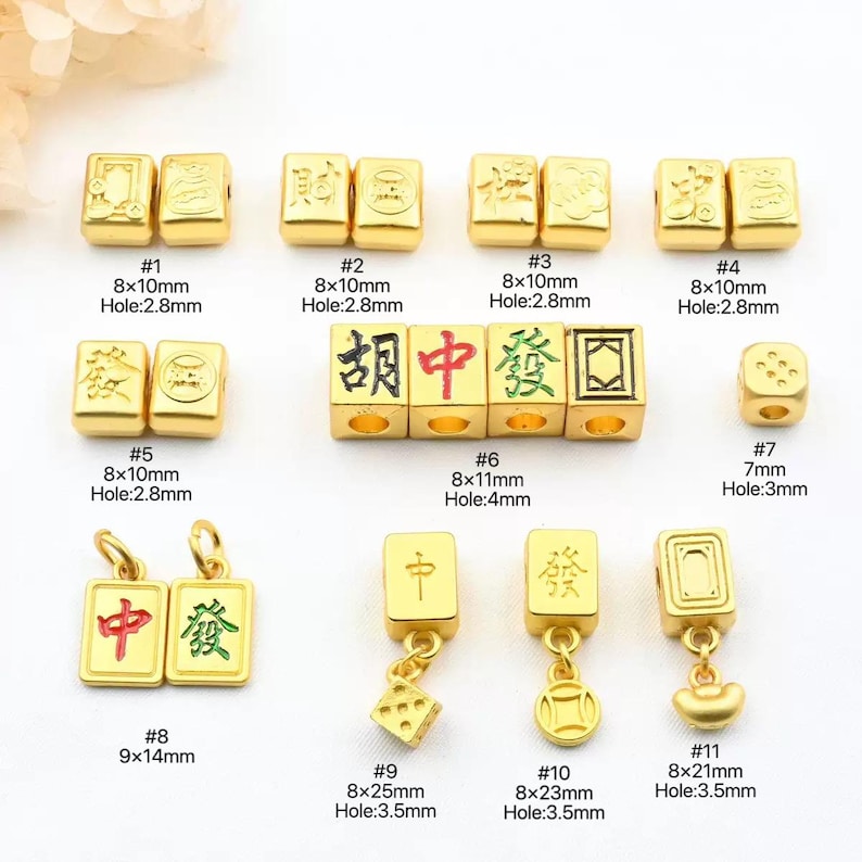 May include: A collection of gold-colored Mahjong-themed charms and beads. The image displays various shapes and sizes, including rectangular beads with Chinese characters, dice-shaped beads, and pendants. Measurements are provided in millimeters.