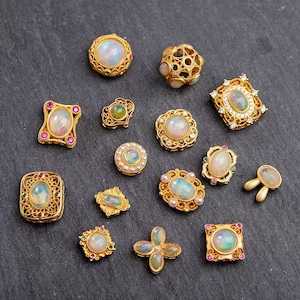 Puede incluir: Una colección de joyas doradas con piedras opalescentes. Los diseños varían, incluyendo formas redondas, cuadradas y florales, algunas con acentos de perlas o piedras preciosas. Las piezas se exhiben sobre una superficie gris oscuro.