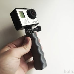 Può includere: Una fotocamera GoPro HERO3 nera montata su un'impugnatura nera con una superficie testurizzata.