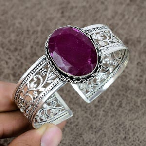 Peut inclure: Un bracelet jonc en argent avec un motif floral orné et une grande pierre précieuse ovale de couleur violet foncé.