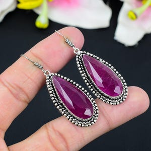 May include: A pair of silver-toned drop earrings featuring large, faceted, teardrop-shaped, deep red gemstones. The gemstones are set in a beaded silver frame with a decorative hook.