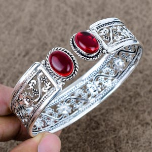 Op de afbeelding: Zilverkleurige manchetarmband met twee ovale, dieprode edelstenen. De armband heeft ingewikkelde bloemen- en krulwerkontwerpen. De edelstenen zijn gezet in een bezel-zetting.