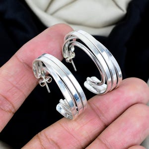 May include: A pair of silver hoop earrings. Each earring features a layered design with three parallel bands. The earrings have a polished finish and are shown against a neutral background. The earrings are held in a person's hand.