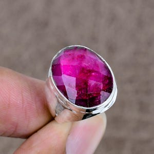 Può includere: Un anello d'argento con una grande gemma ovale sfaccettata di colore magenta vibrante. La gemma è il punto focale, catturando la luce con le sue molteplici sfaccettature. La fascia dell'anello è in argento lucido.