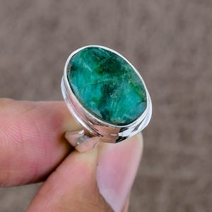 Puede incluir: Un anillo de plata con una gran gema verde ovalada. La gema tiene una superficie texturizada con diferentes tonos de verde. La banda del anillo es de plata pulida.