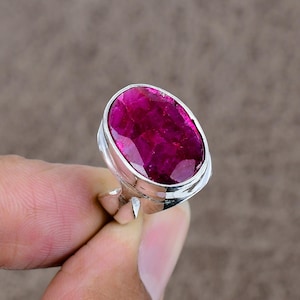 May include: A silver ring featuring a large, oval-cut ruby gemstone. The ruby is a vibrant shade of pink-red, set in a polished silver band. The ring's design is simple, highlighting the gemstone's color and shape.