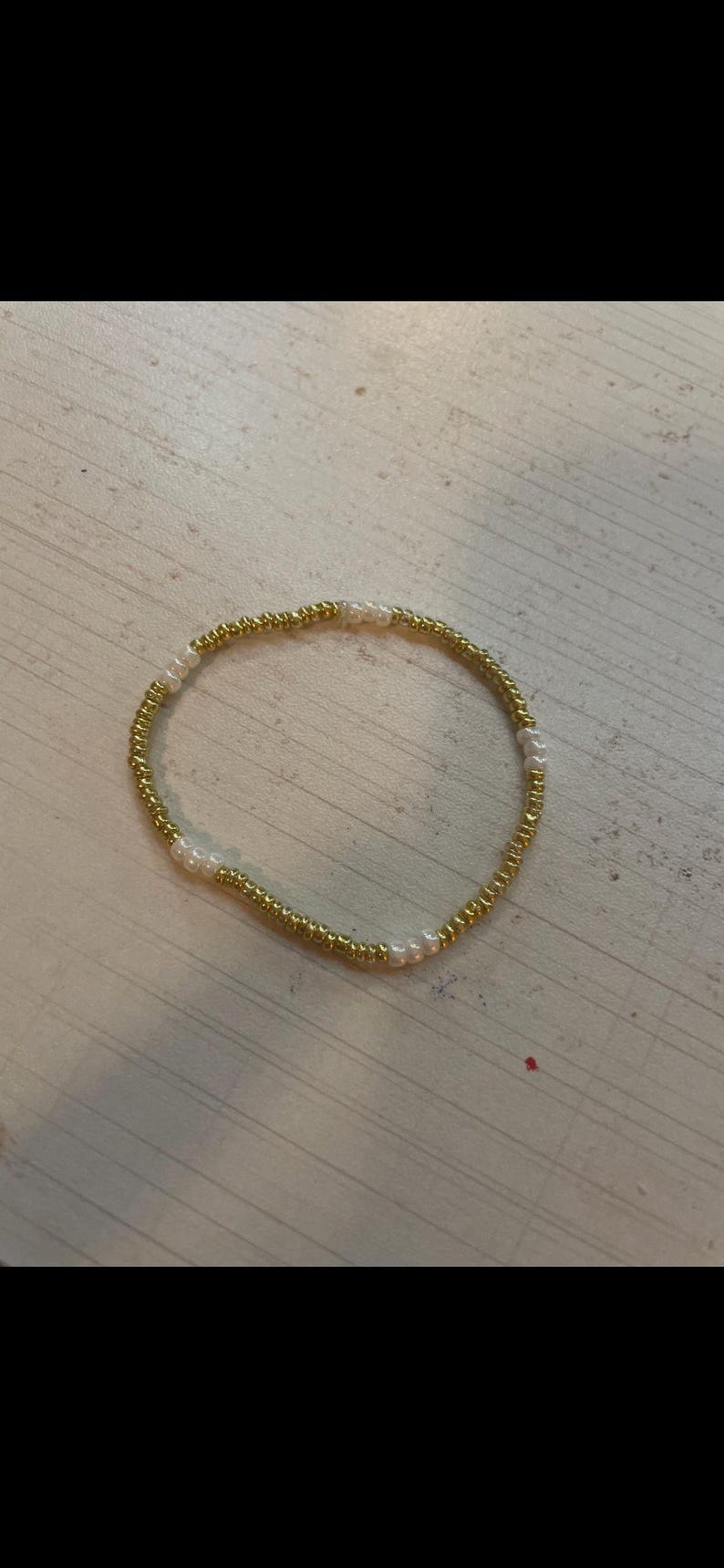 May include: A gold beaded bracelet with small, round, white beads interspersed throughout. The bracelet is circular and sits on a light-coloured surface. The beads are uniform in size and shape.
