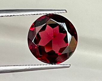 May include: A deep red gemstone with a faceted cut, held by tweezers against a white background.