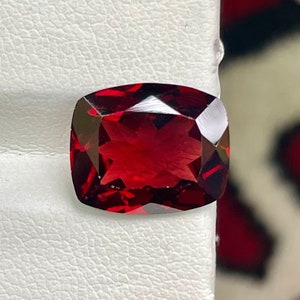 May include: A deep red gemstone with a cushion cut, displayed on a white surface.