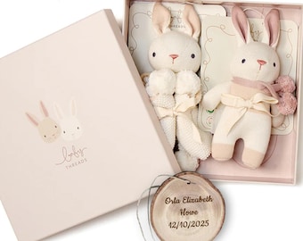 May include: A gift box with two plush bunny toys. The bunnies are cream-colored with pink accents and bows. The box is light pink with a bunny illustration and the text "baby threads". A wooden tag with engraved text is included.