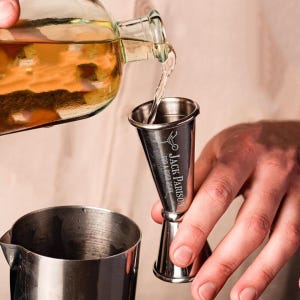 May include: A stainless steel jigger with a conical shape and a measurement line. The jigger is engraved with the text "Jack Parison for a good drink". A hand is pouring liquid from a glass bottle into the jigger.