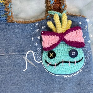 Pu&ograve; includere: Un personaggio Stitch in crochet blu turchese con un fiocco rosa e viola, capelli gialli e occhi neri. Il personaggio &egrave; attaccato a una borsa di jeans con un ago e filo bianchi.